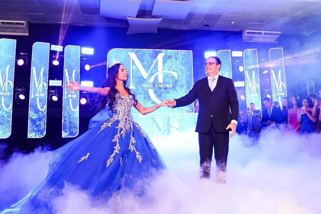 Entrada da debutante de mãos dadas com o pai, em vestido azul, sob fumaça baixa — cerimônia de 15 anos em Brasília