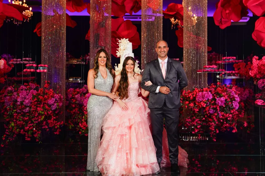 Debutante em vestido rosa entre a mãe e o pai, diante da decoração de flores — festa de 15 anos em Brasília