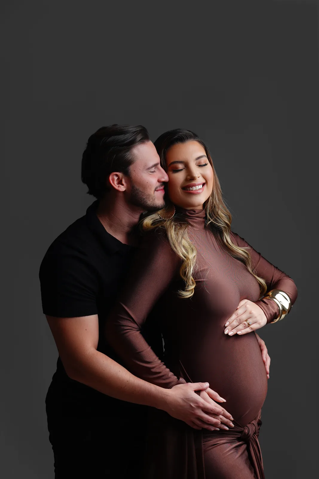 Casal grávido, ele beijando a testa dela, vestido marrom, mão na barriga