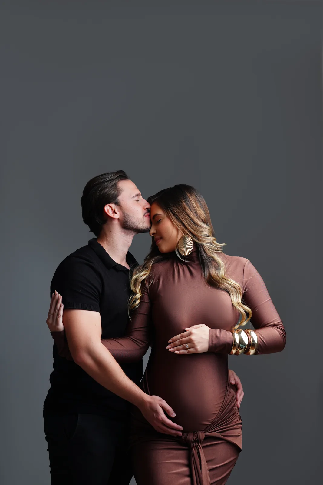 Casal grávido, ele beijando a testa dela, pulseiras douradas, vestido marrom