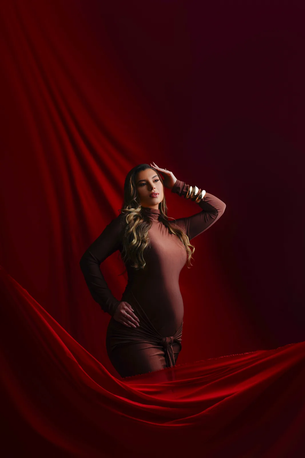 Gestante em vestido vinho, fundo de cortina vermelha, mão na cabeça