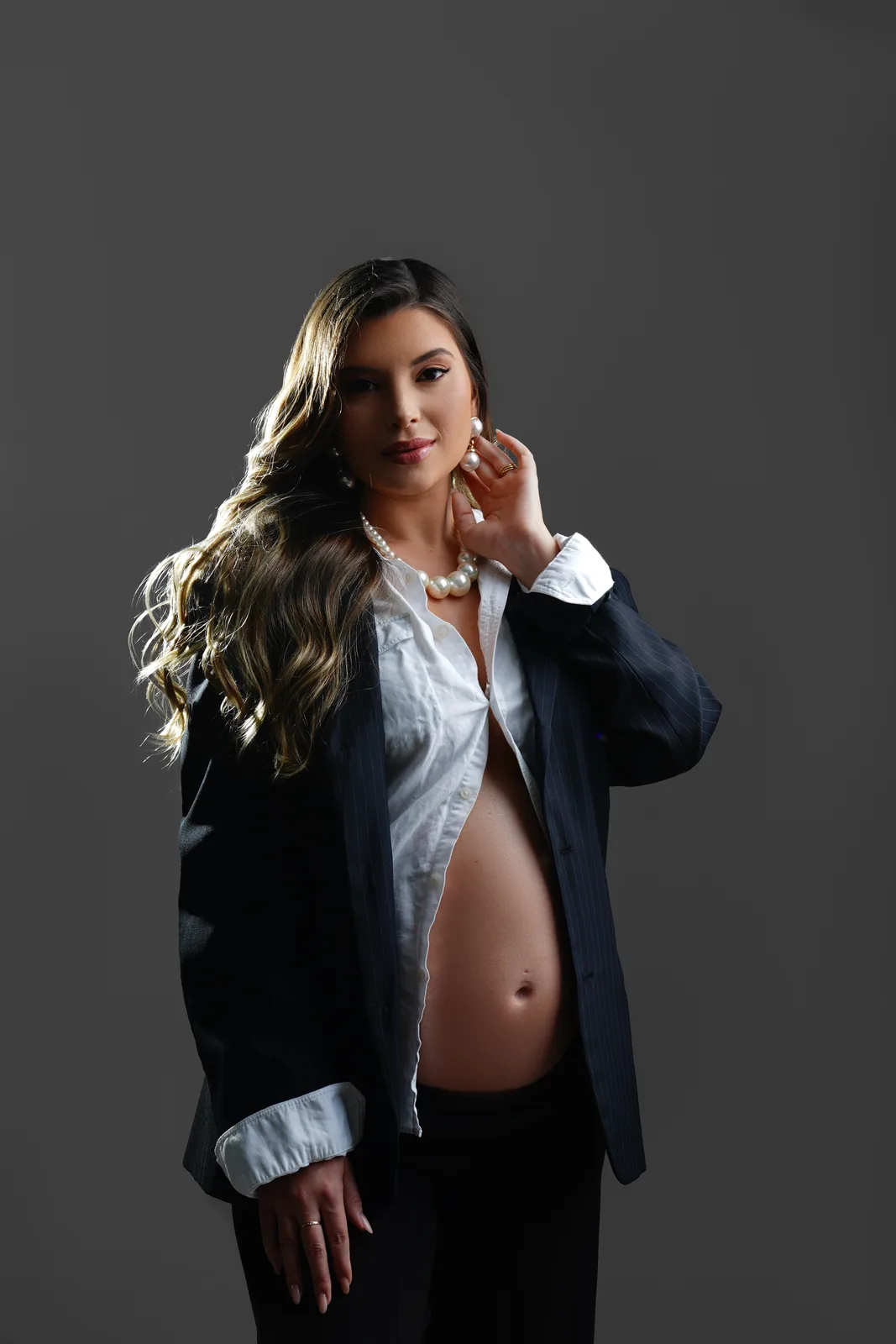 Gestante em blazer e camisa branca aberta, colar de pérolas, barriga à mostra