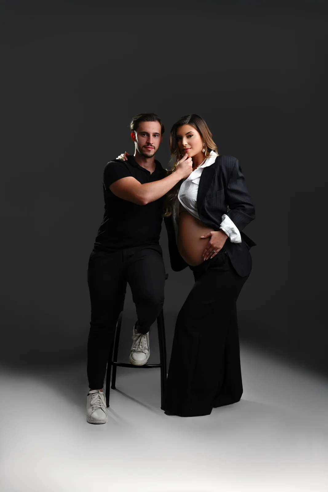 Casal grávido, ele sentado em banqueta, blazer preto e calça pantalona