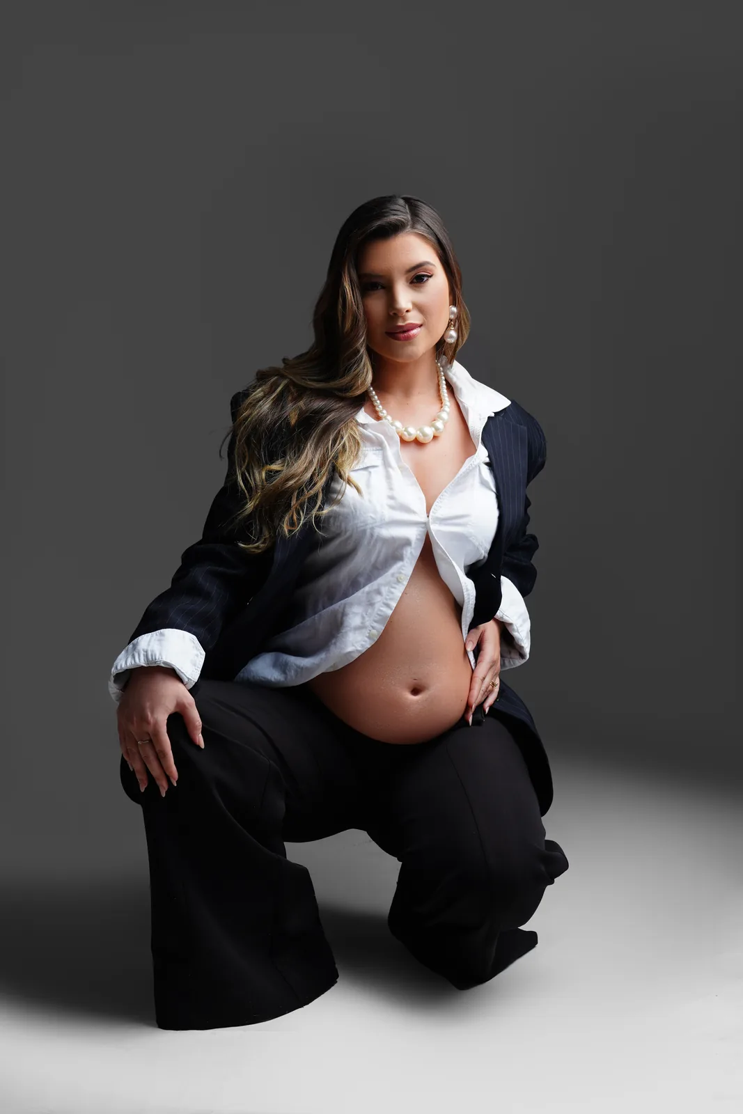 Gestante ajoelhada, blazer aberto, colar de pérolas, calça pantalona preta