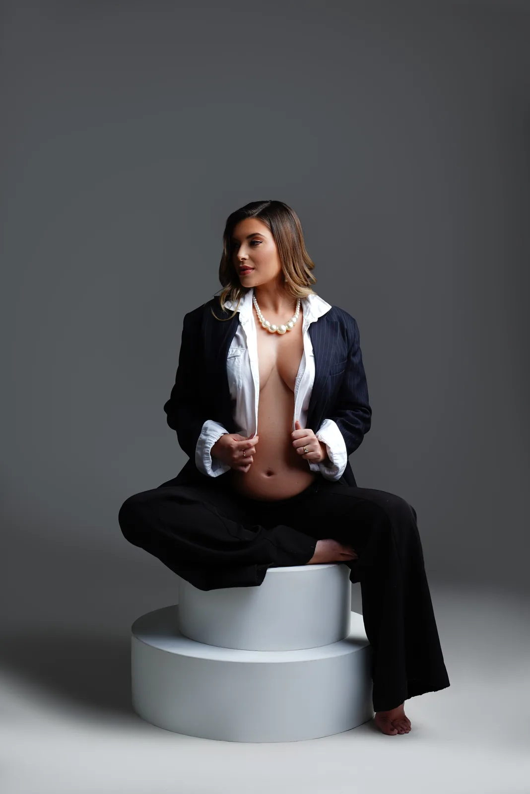 Gestante sentada em cubo branco, blazer e colar de pérolas, corpo inteiro