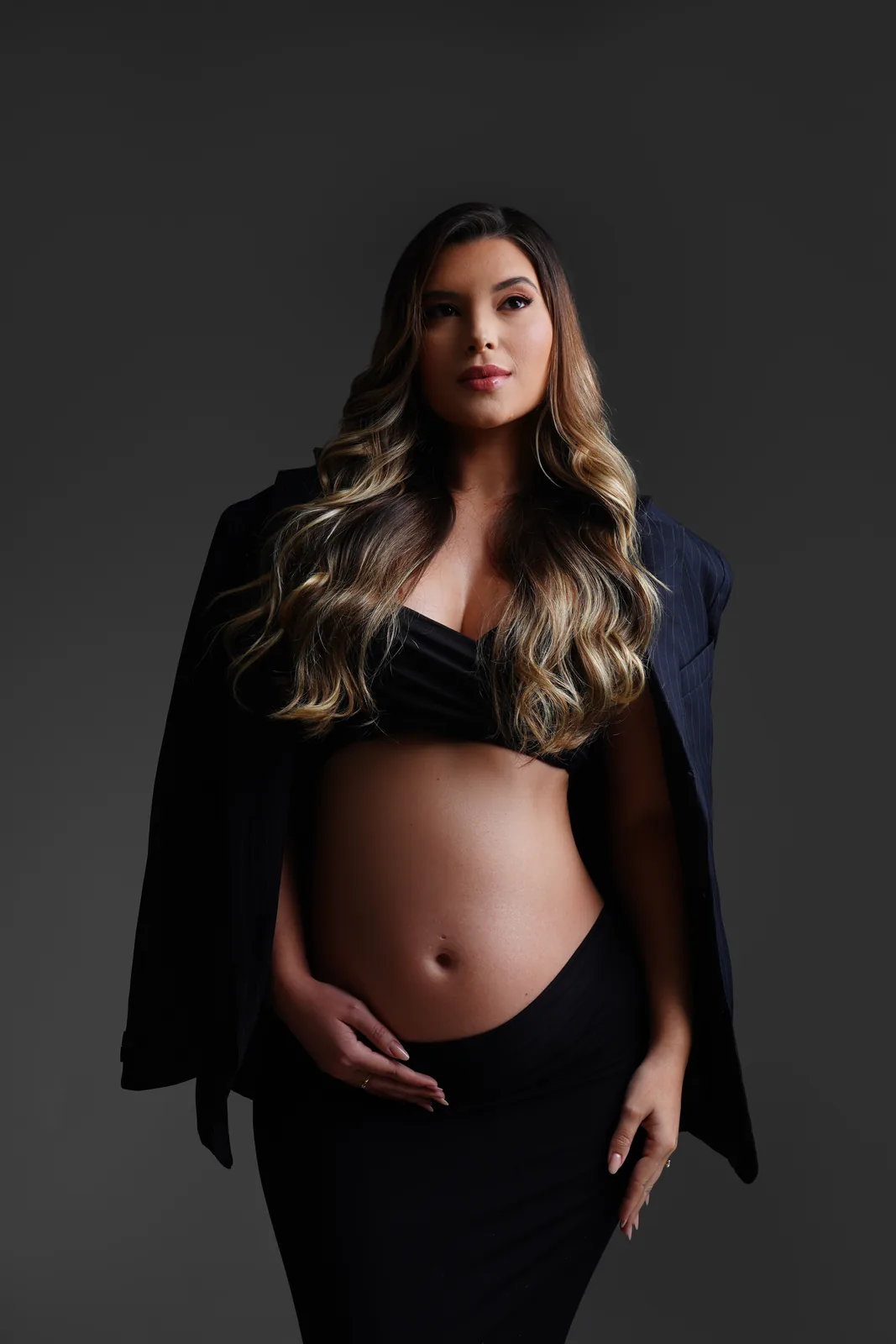 Gestante em top preto e blazer aberto, barriga à mostra, mão na cintura