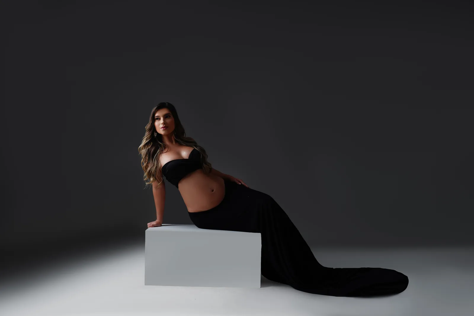 Gestante sentada em cubo branco, top preto e saia longa, corpo inteiro