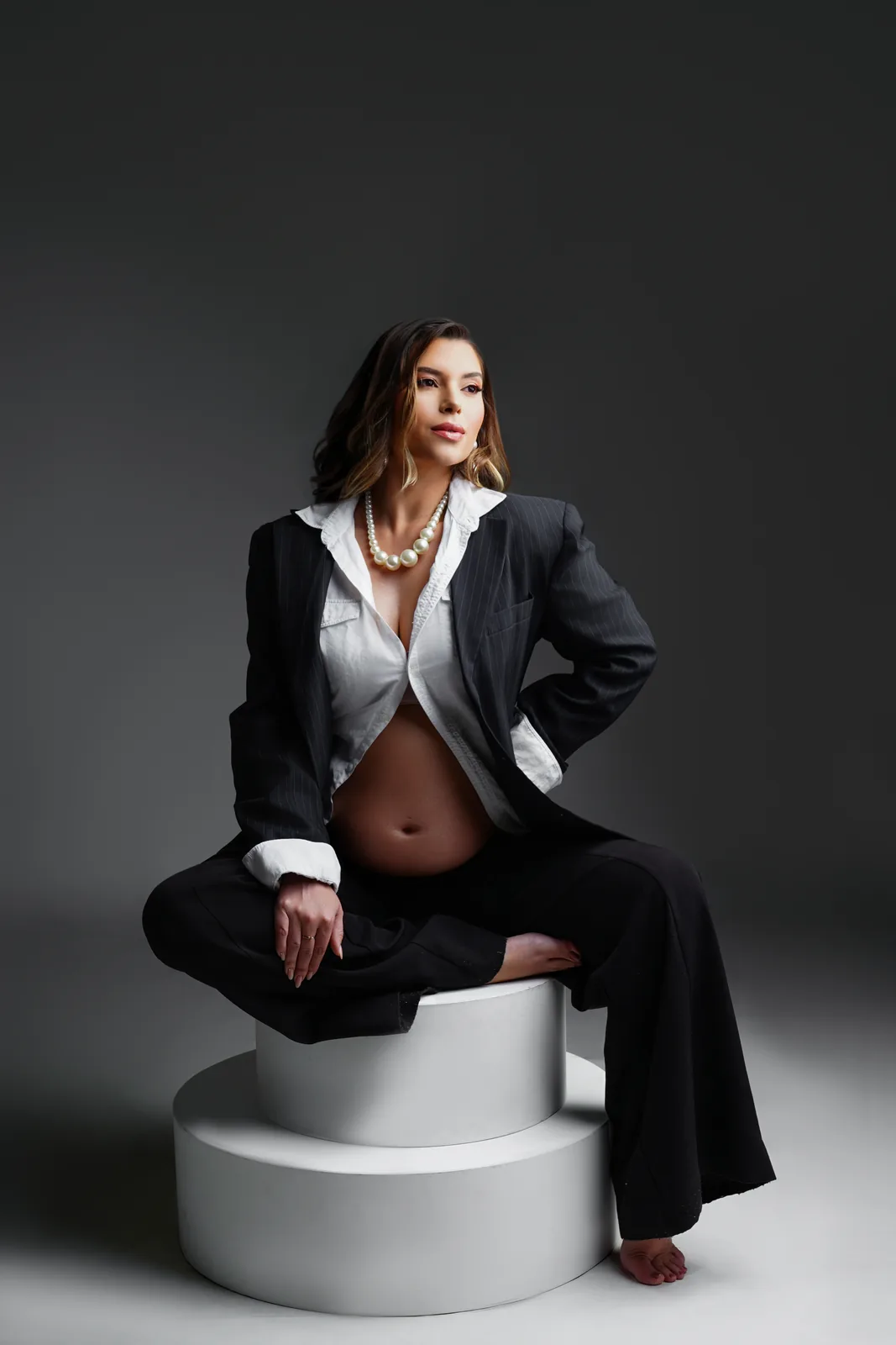 Gestante sentada em pedestal, blazer e colar de pérolas, calça preta, corpo inteiro