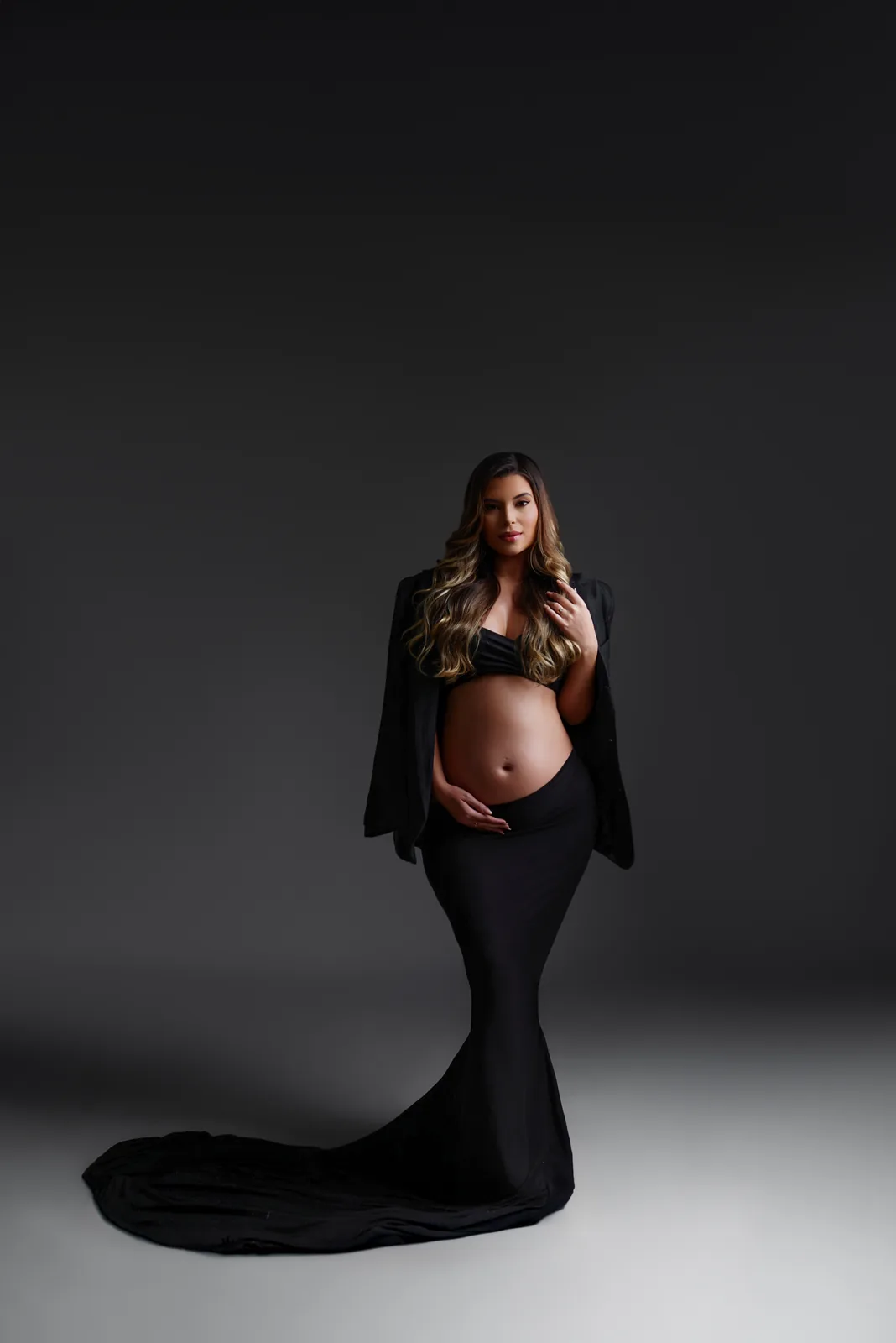 Gestante em vestido preto sereia com blazer sobre os ombros, mão na barriga