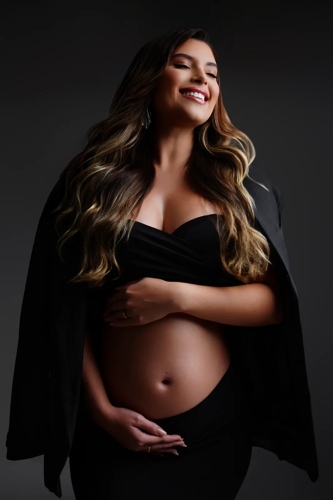 Gestante em top preto e blazer, mão cruzada na barriga, sorriso aberto