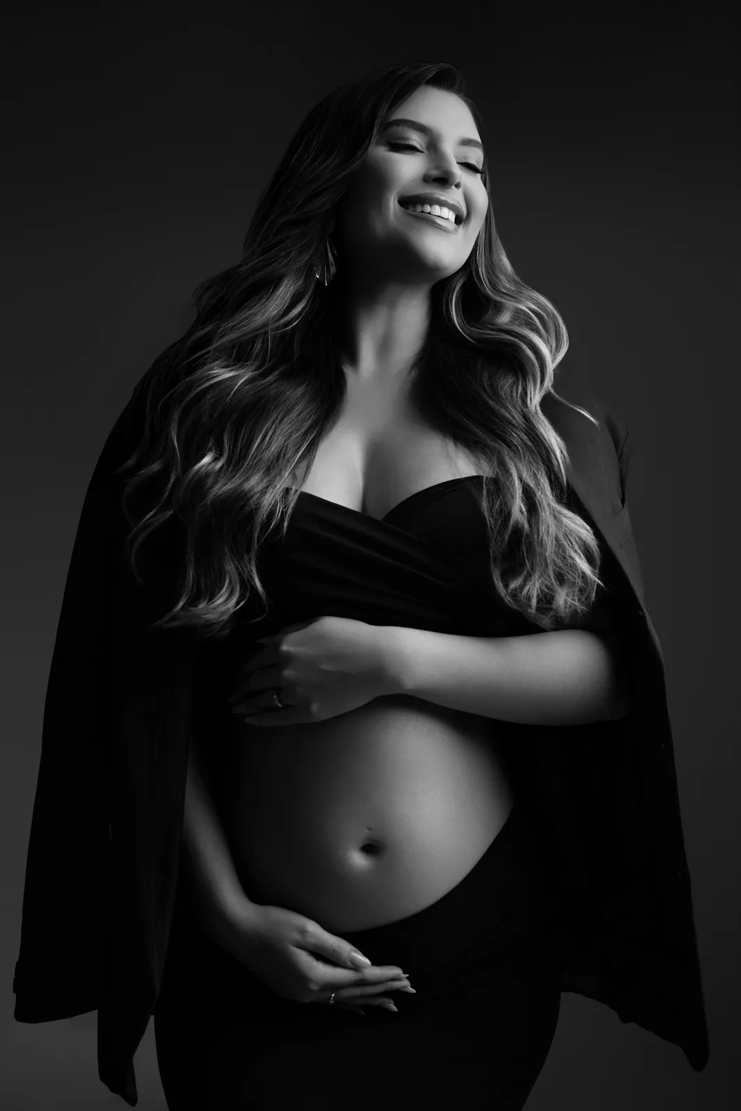 Gestante em top preto e blazer, sorriso aberto, mesma cena em preto e branco