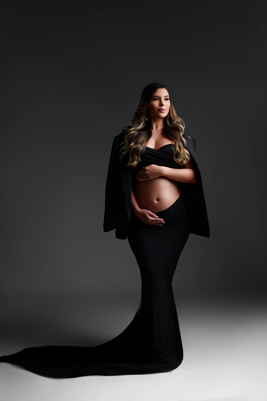 Gestante em top preto e blazer, mão na barriga, olhando de lado