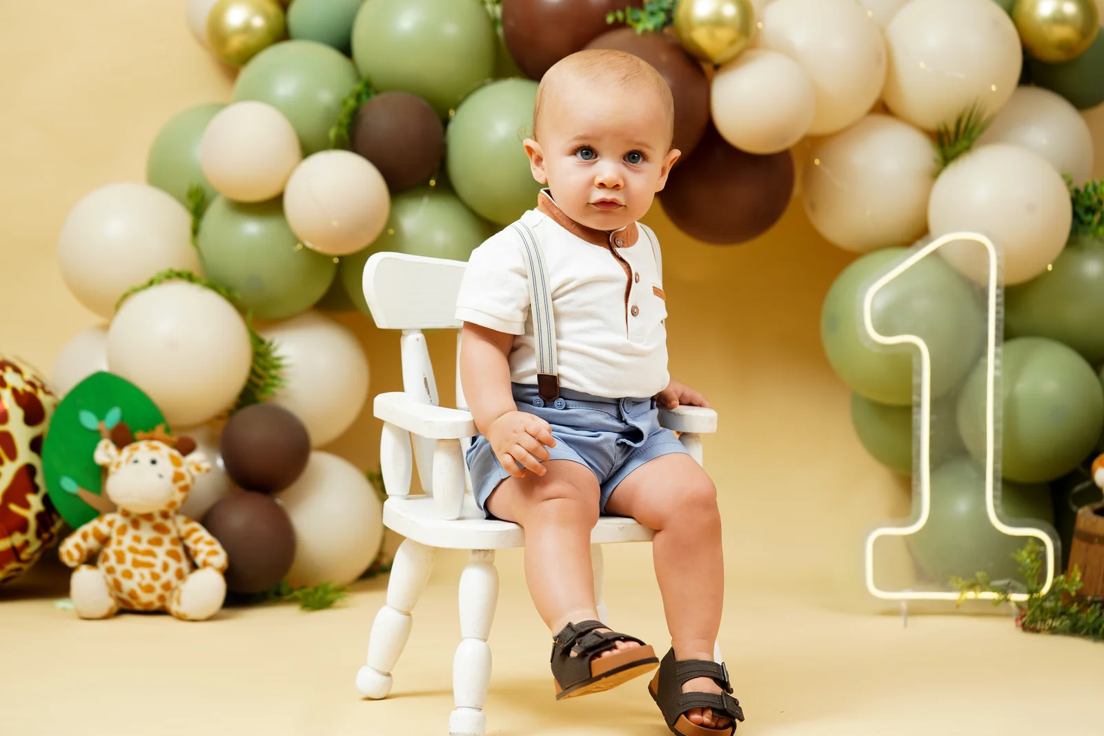 Bebê sentado na cadeirinha branca ao lado do número 1 dourado, decoração de balões verde e marrom — ensaio smash the cake