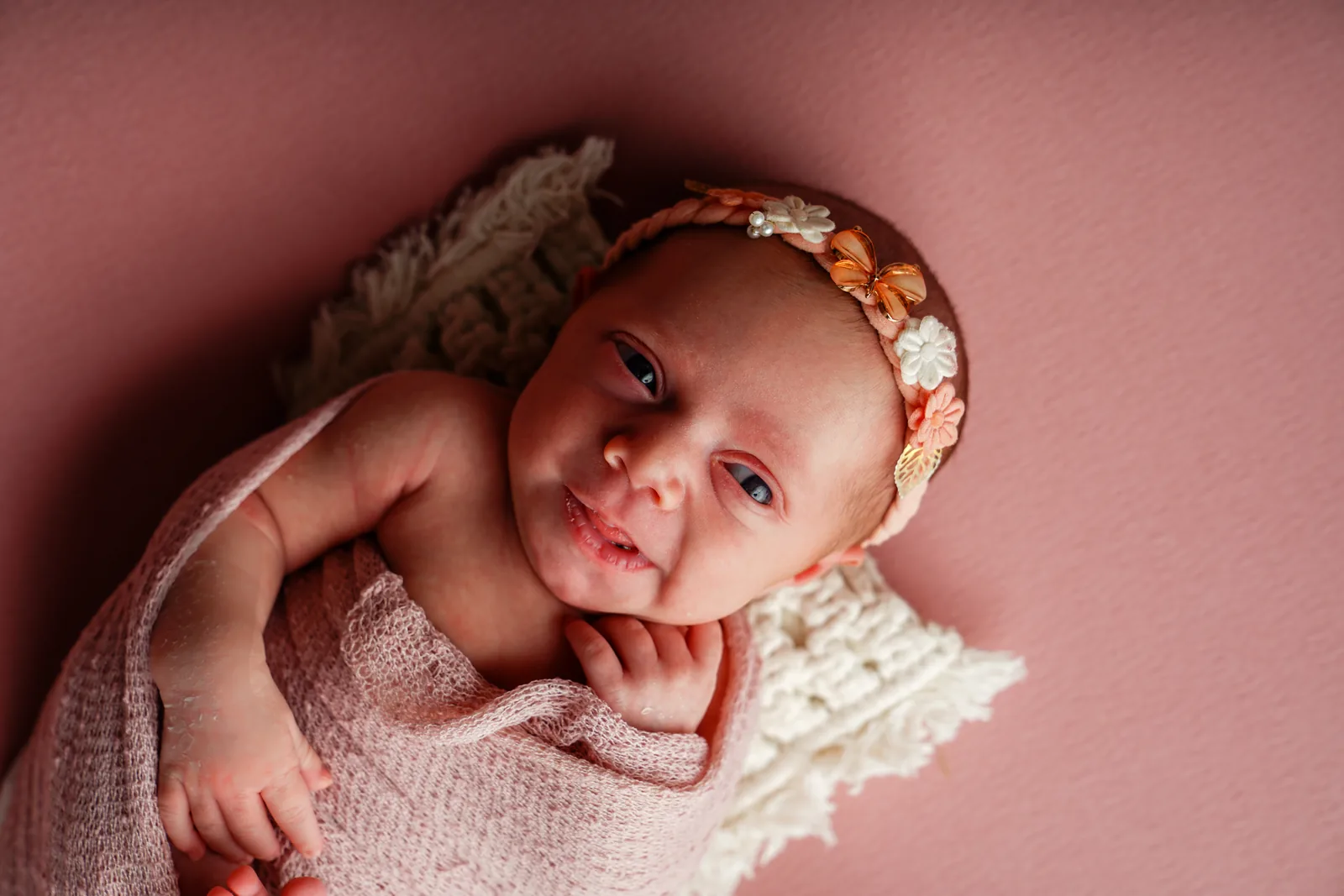 Recém-nascida com olhos abertos e sorriso discreto, faixa floral na cabeça, manta rosa
