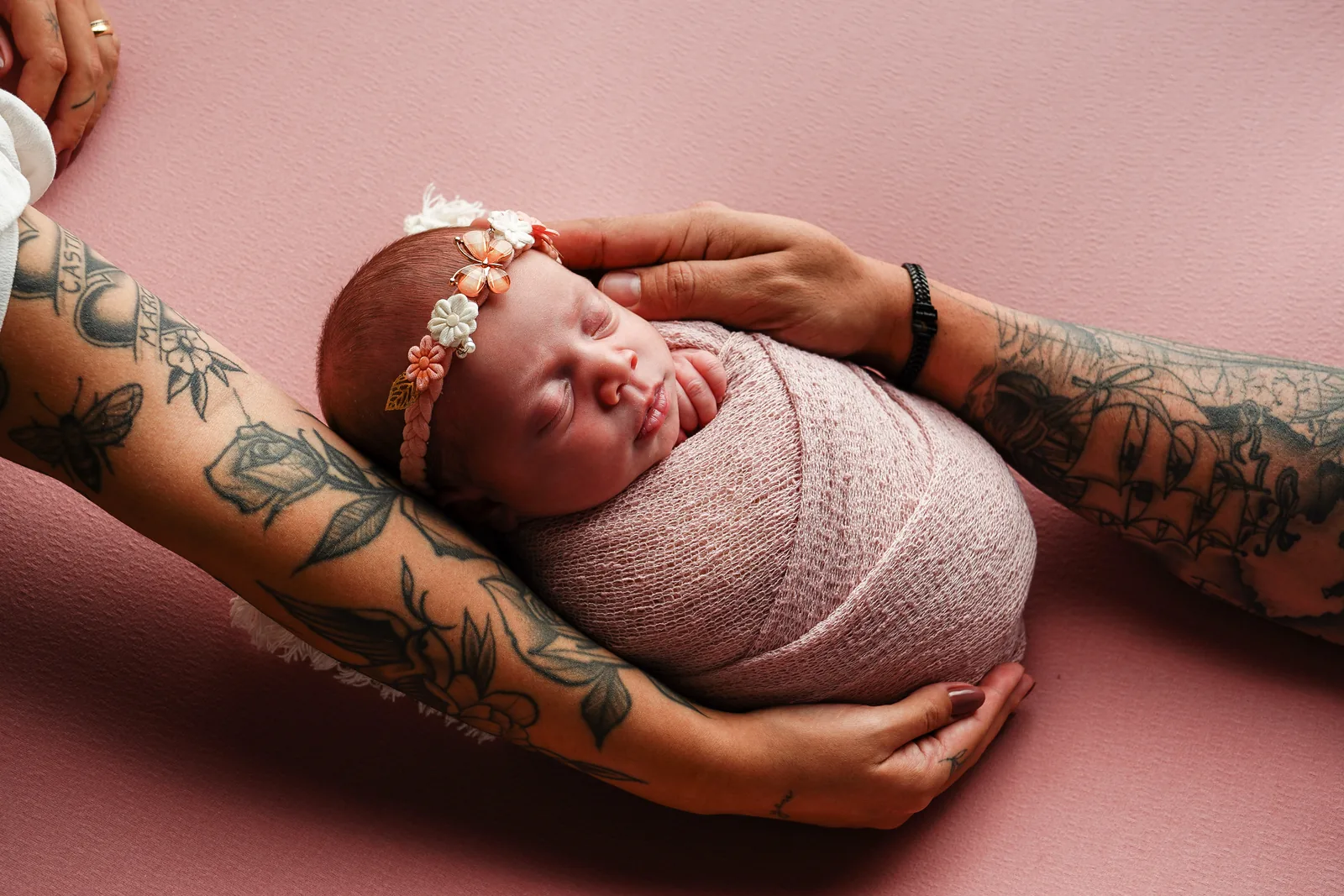 Recém-nascida enrolada em manta rosa, apoiada nos braços tatuados dos pais, fundo rosa