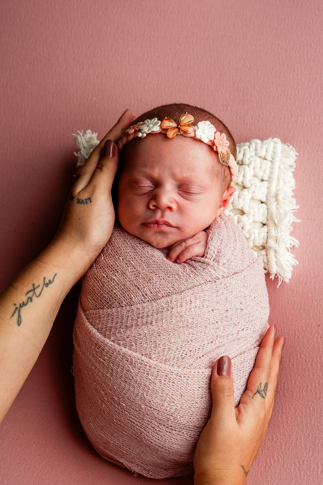 Recém-nascida dormindo, faixa floral na cabeça, mão da mãe tocando sua cabeça com carinho