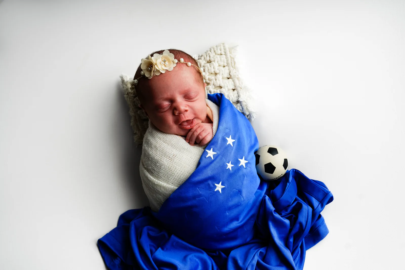 Recém-nascida enrolada em camisa de time de futebol azul com estrelas, bolinha de futebol ao lado