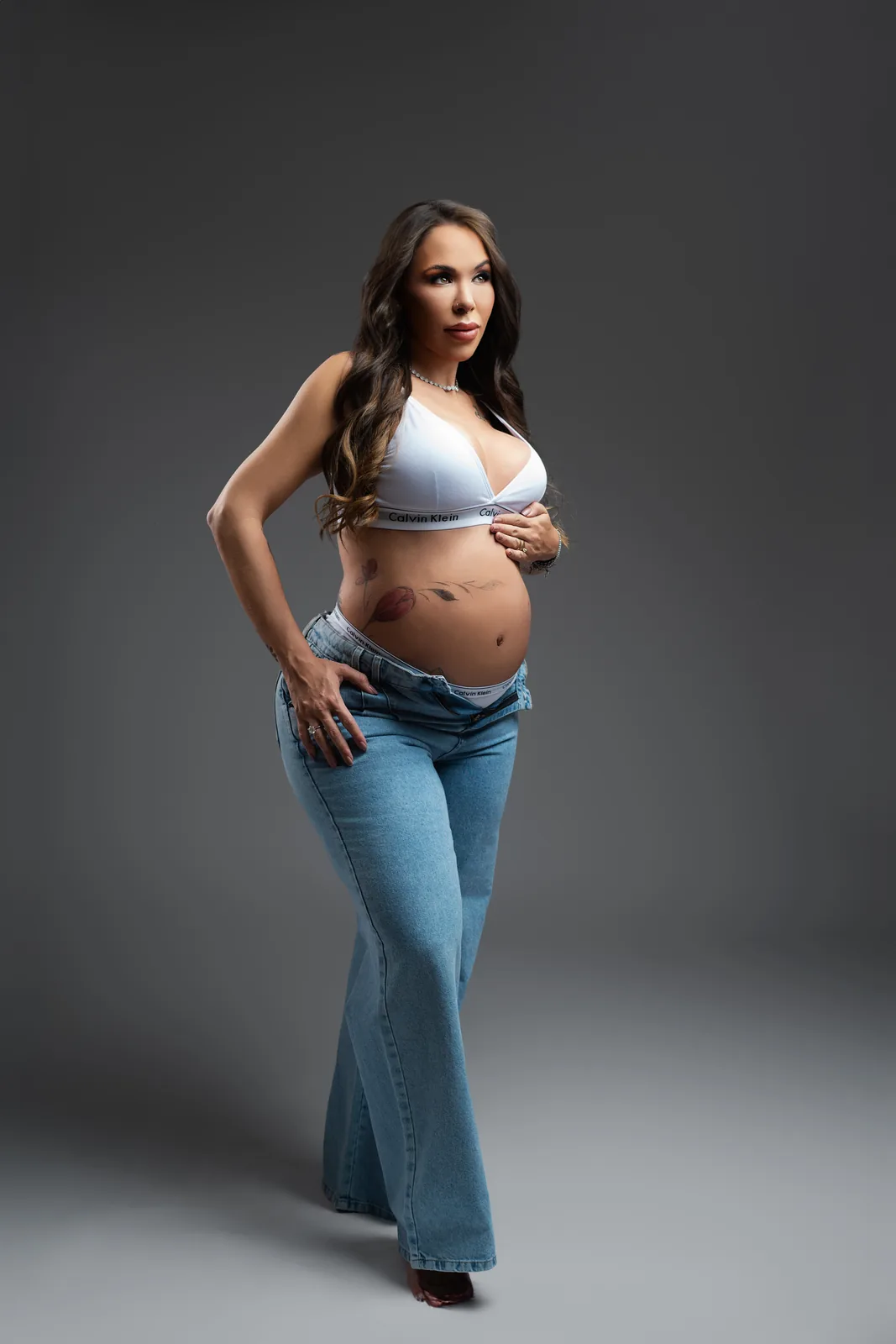 Gestante em top branco Calvin Klein e jeans, mão na cintura, olhando de lado — ensaio gestante em estúdio