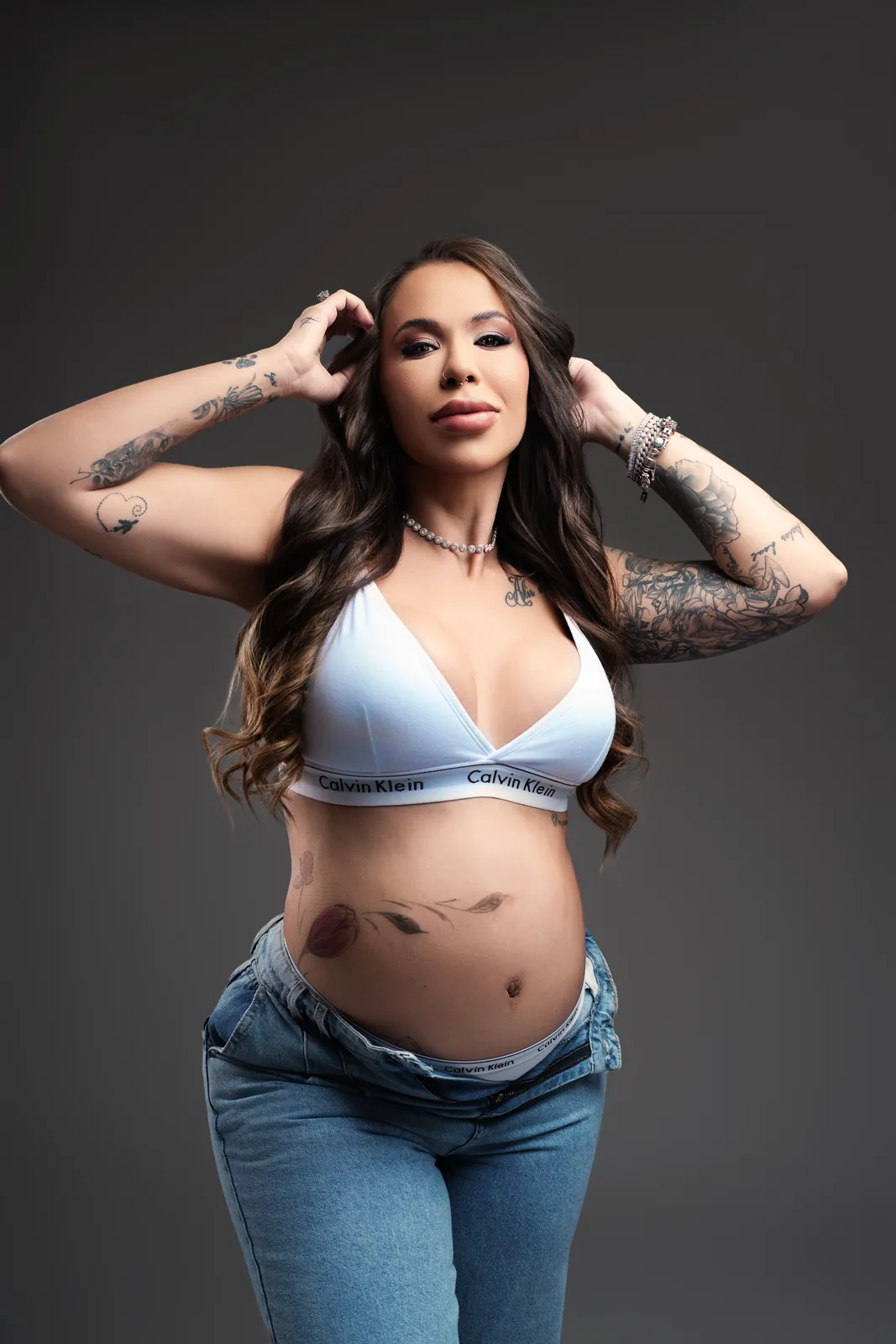 Gestante em top branco Calvin Klein, mãos no cabelo, olhando para a câmera — ensaio gestante em estúdio