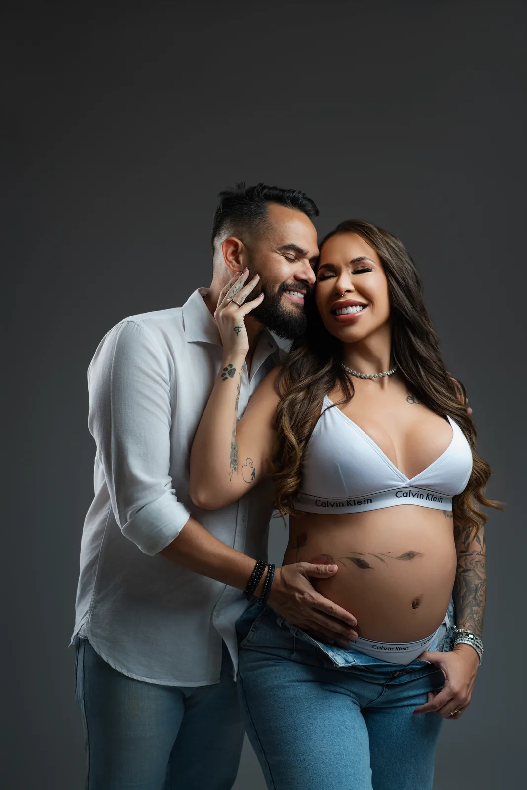 Gestante em top branco Calvin Klein e jeans, mão na cabeça, corpo inteiro — ensaio gestante em estúdio