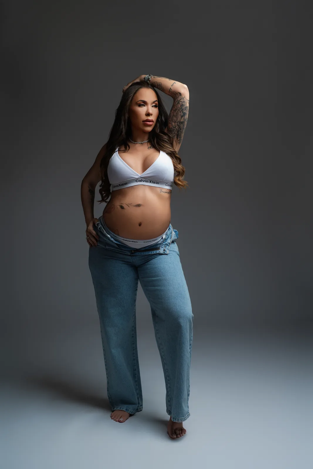 Gestante em top branco Calvin Klein e jeans, mão na cintura, tatuagens no braço à mostra