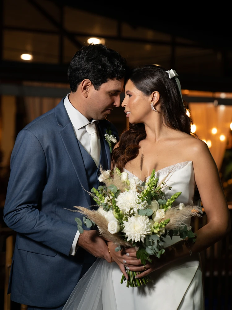 Noivo encostando a testa na noiva, os dois sorrindo próximos, ela com bouquet de flores brancas — fotografia de casamento à noite em Brasília