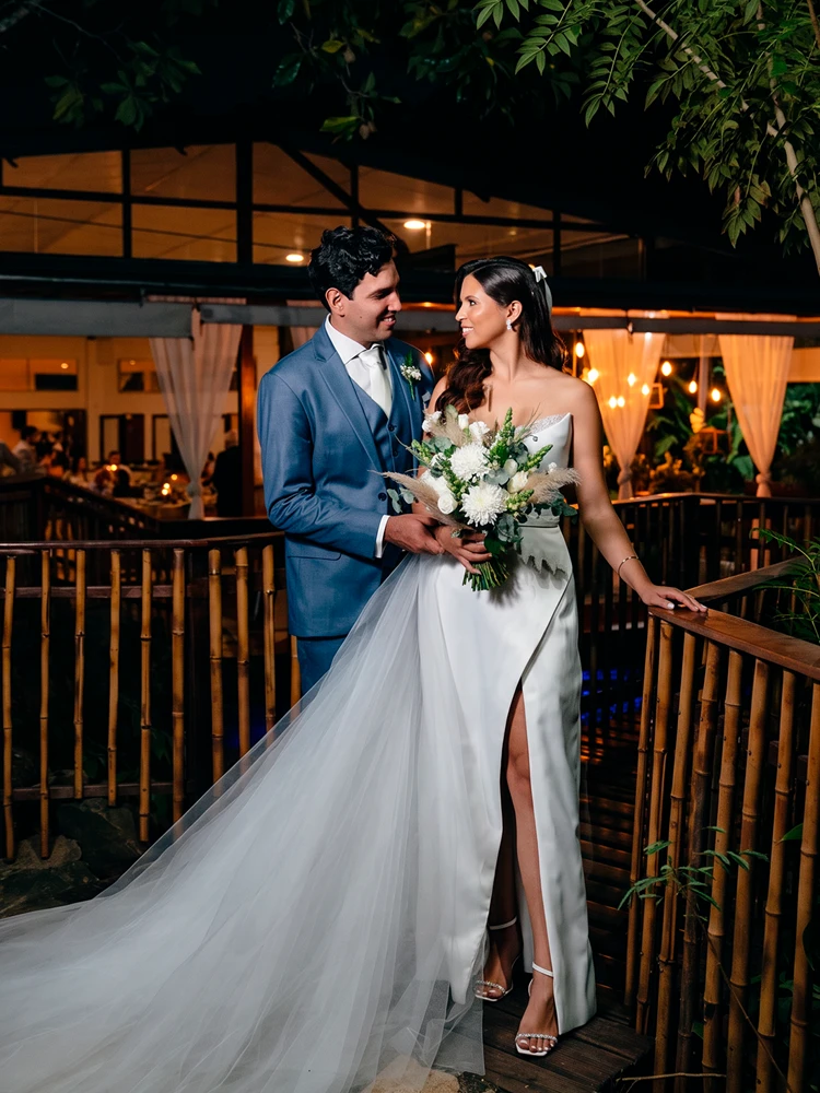 Noivos sorridentes num deck de madeira iluminado, ela em vestido com fenda e véu curto — fotografia de casamento em Brasília