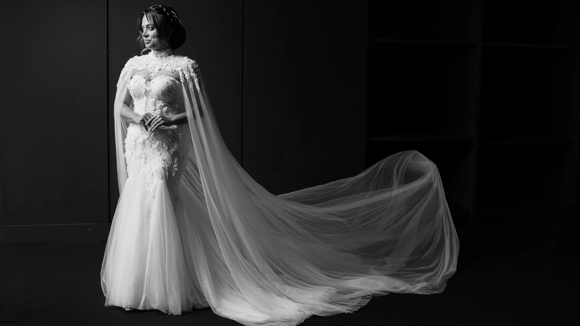Noiva de vestido sereia e capa bordada, olhar de perfil em estúdio escuro, em preto e branco — retrato editorial de casamento em Brasília