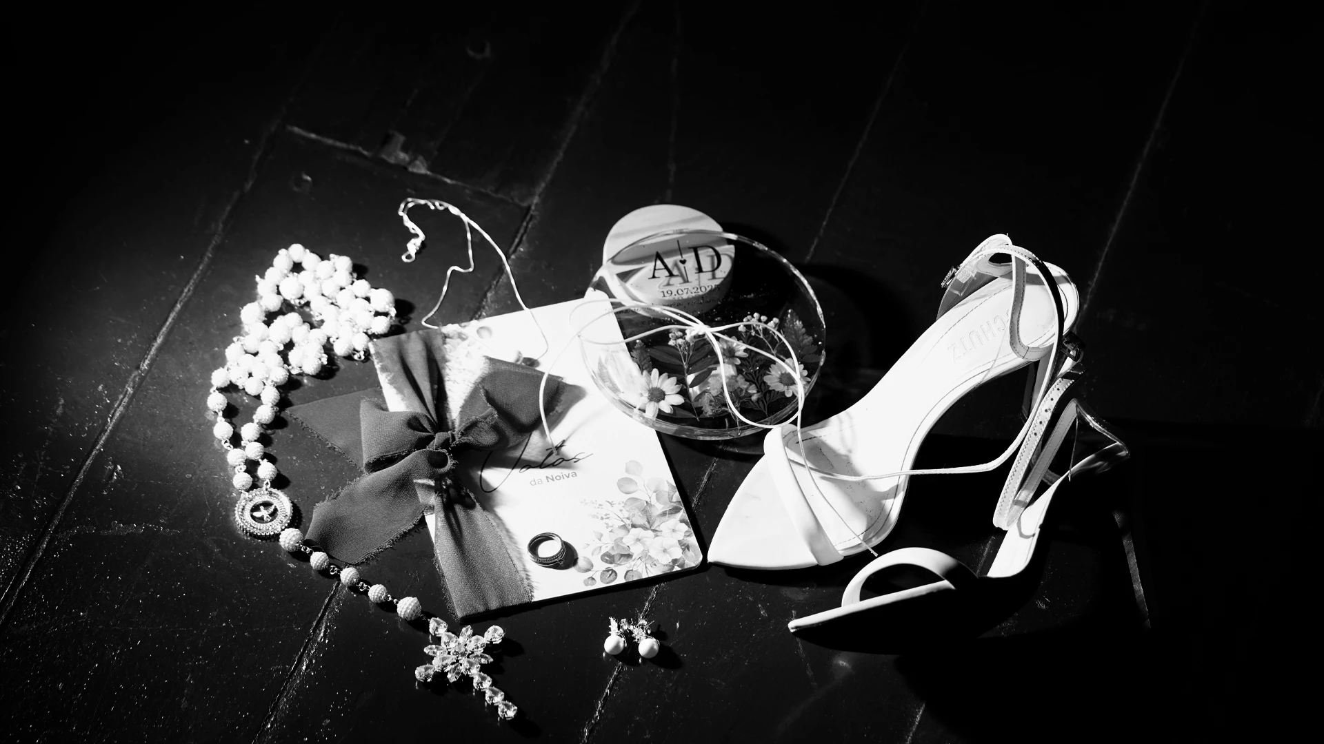 Sapato de noiva, colar de pérolas, aliança e caderno 'Votos da Noiva' sobre piso escuro, em preto e branco — still de casamento em Brasília