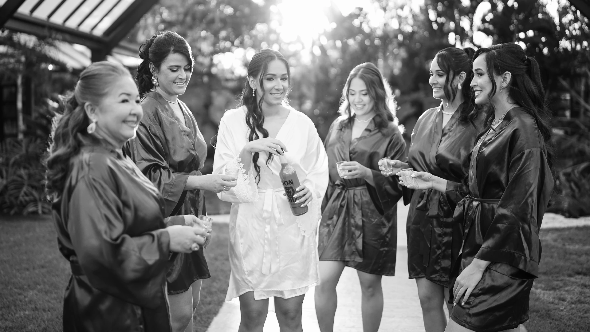 Noiva brindando com tequila ao lado das madrinhas, todos sorrindo sob luz de fim de tarde, em preto e branco — preparativos de casamento em Brasília