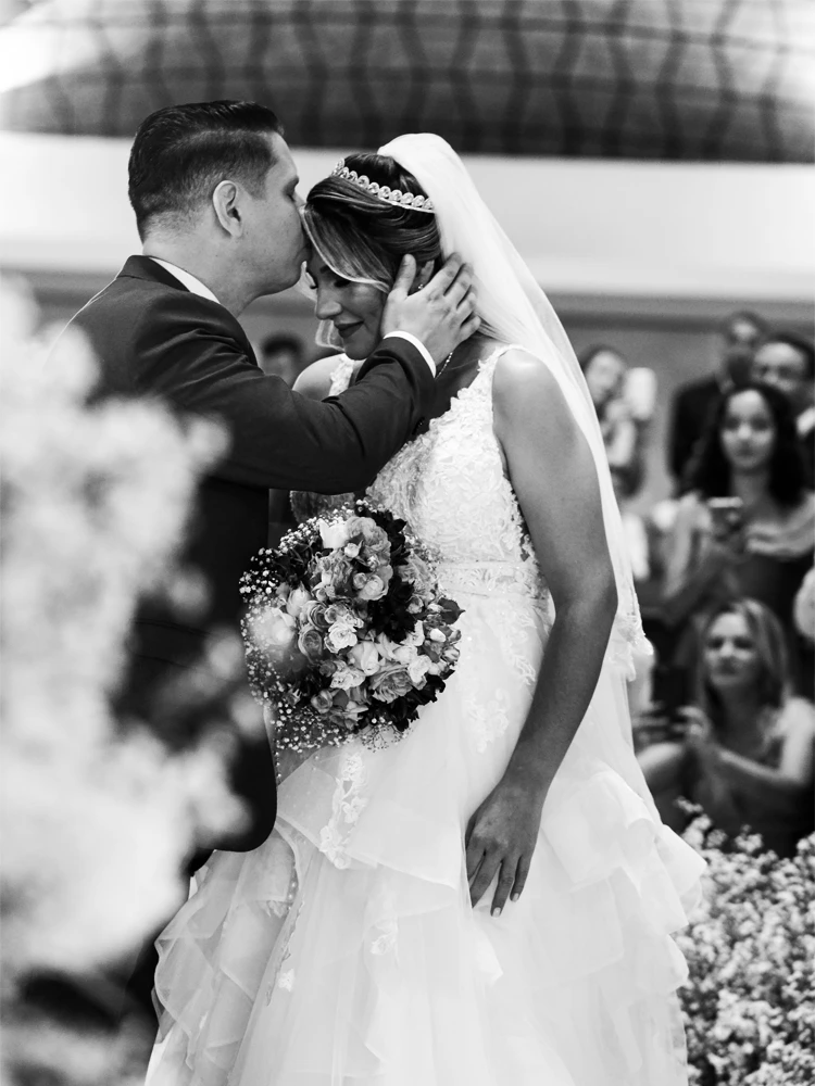 Noivo beijando a testa da noiva de véu e tiara durante a cerimônia, em preto e branco — casamento em Brasília
