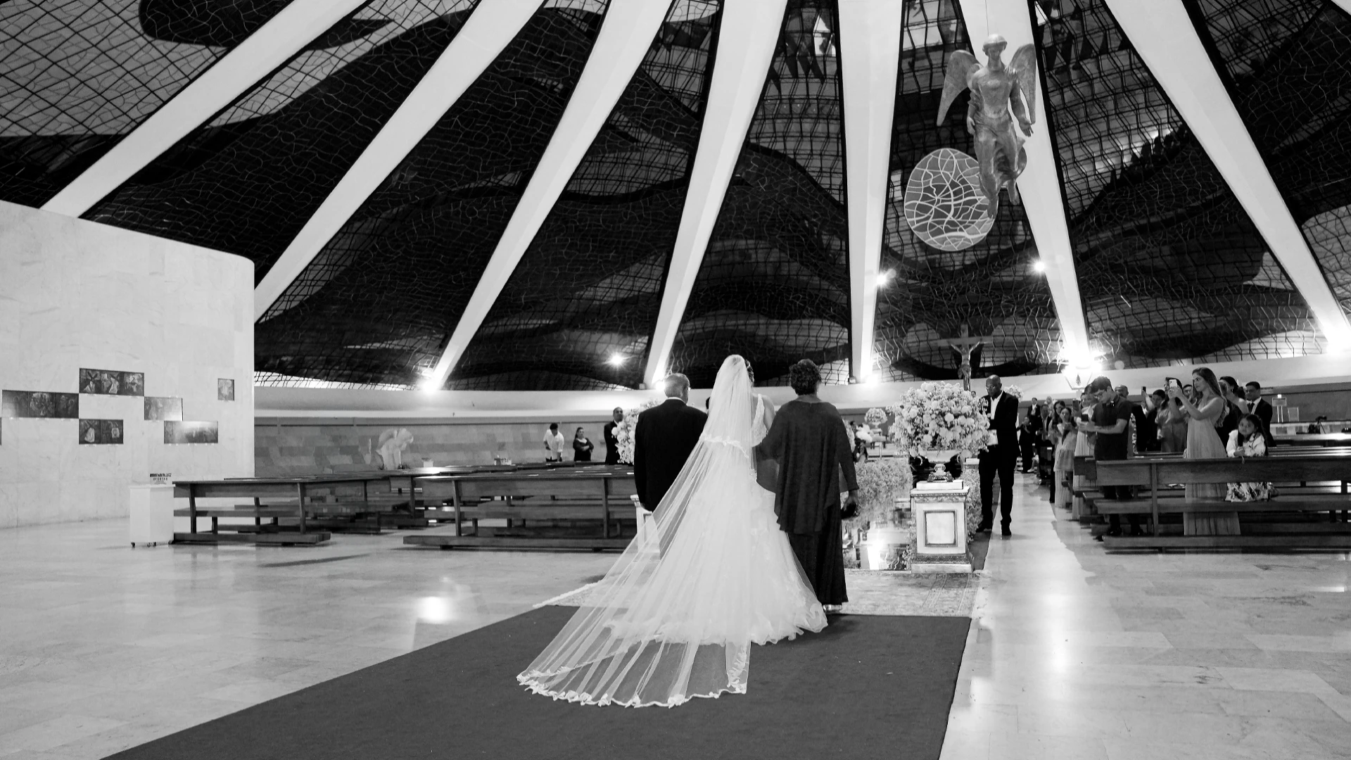 Noiva sendo conduzida pelo pai e pela mãe pelo corredor da Catedral de Brasília, em preto e branco — entrada da noiva no casamento
