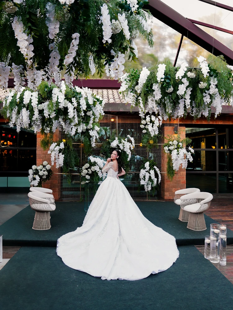 Noiva sob instalação de flores brancas suspensas, sorrindo por cima do ombro — decoração de casamento em Brasília