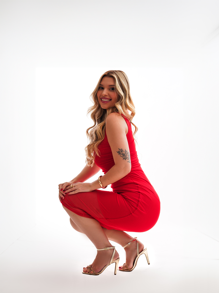 Retrato agachada em vestido vermelho e sandálias douradas, fundo claro — ensaio feminino em Brasília