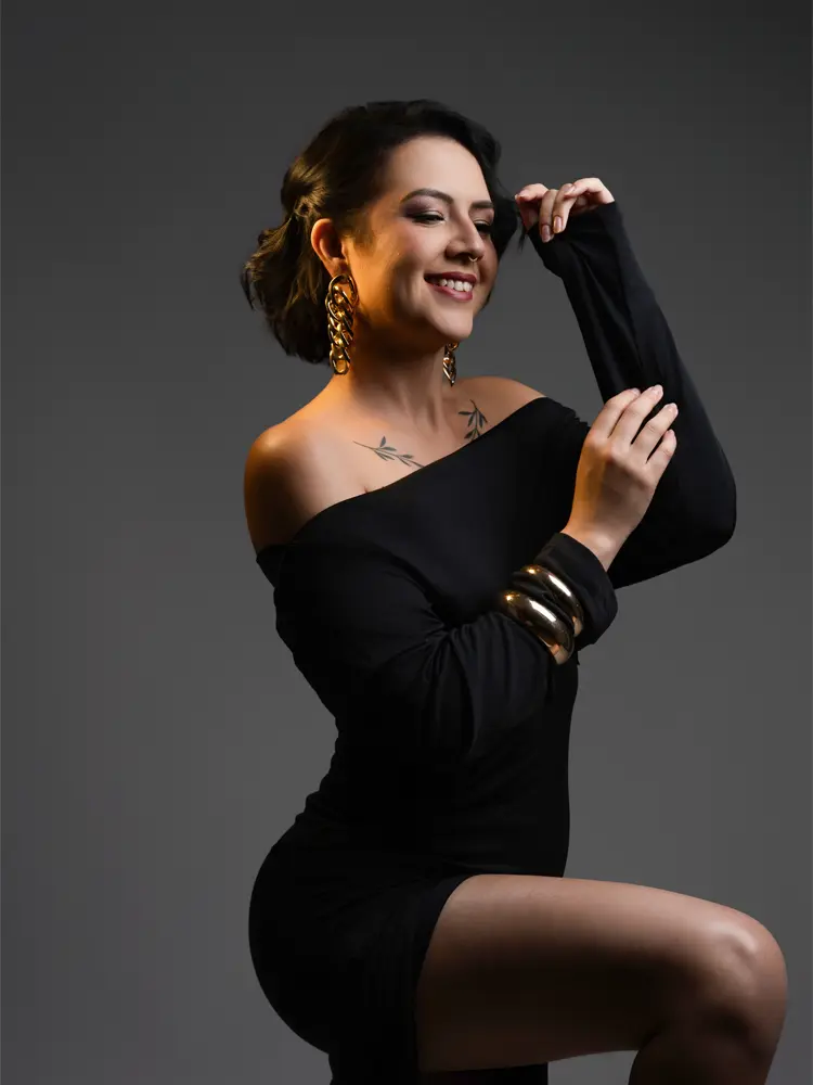 Mulher rindo em vestido preto de ombro só e joias douradas — ensaio feminino em estúdio no Lago Sul, Brasília