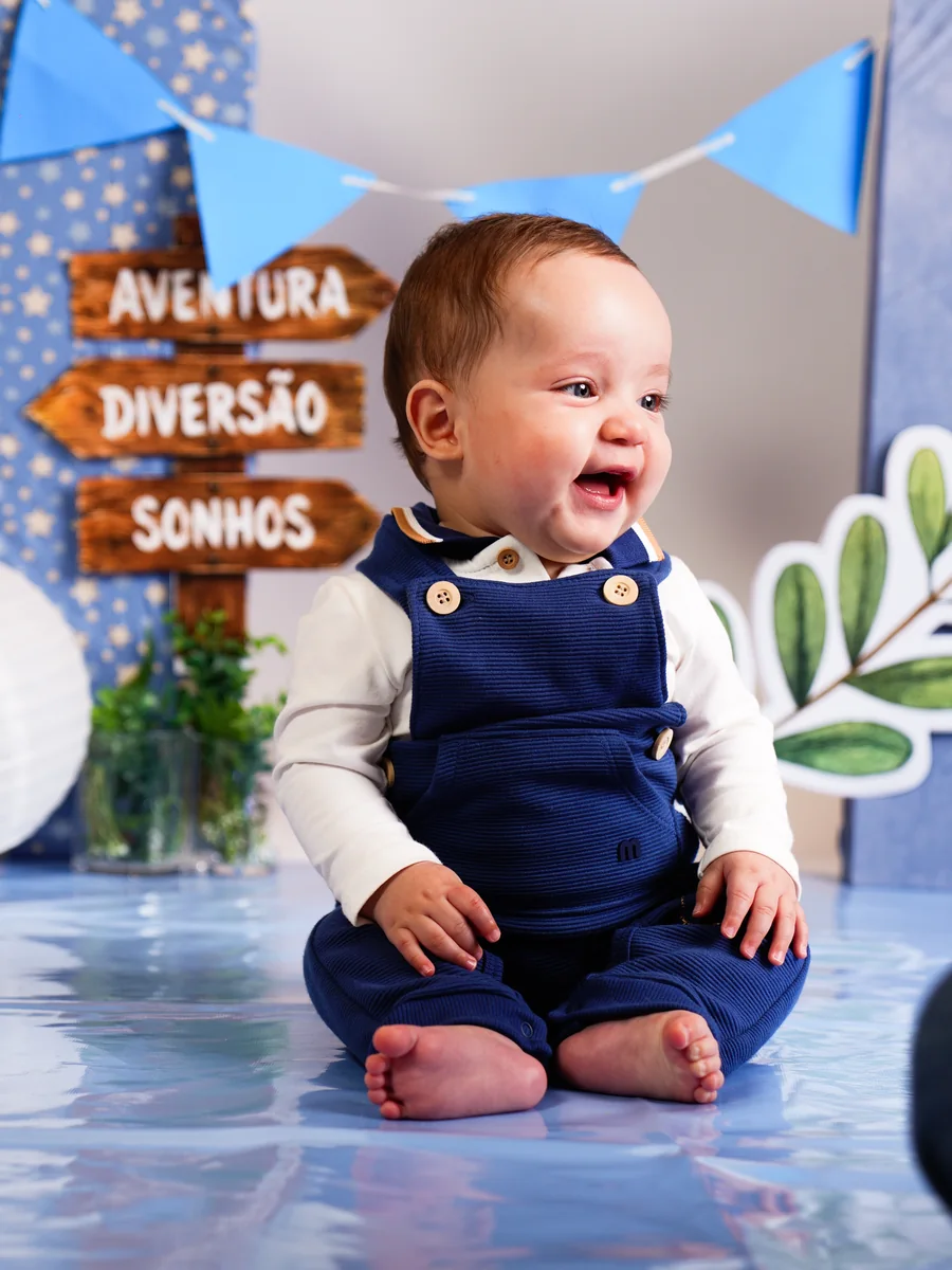 Bebê de macacão azul, sorrindo, sentado diante de placas de madeira “Aventura, Diversão, Sonhos” — ensaio infantil de 6 meses em Brasília