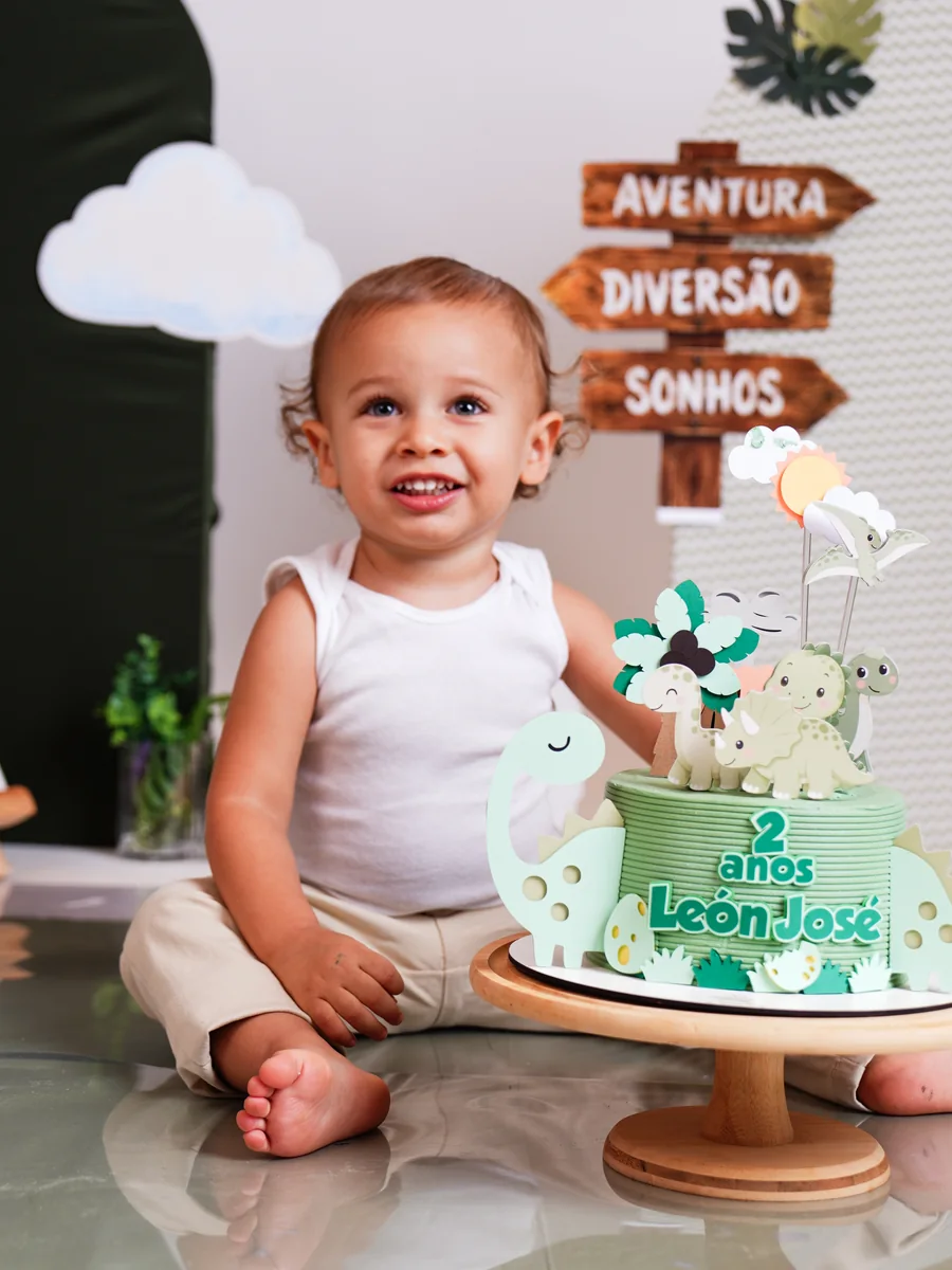 Criança sentada ao lado de um bolo verde de dinossauro com o número 2, diante de placas de madeira “Aventura, Diversão, Sonhos” — Smash the Cake em Brasília