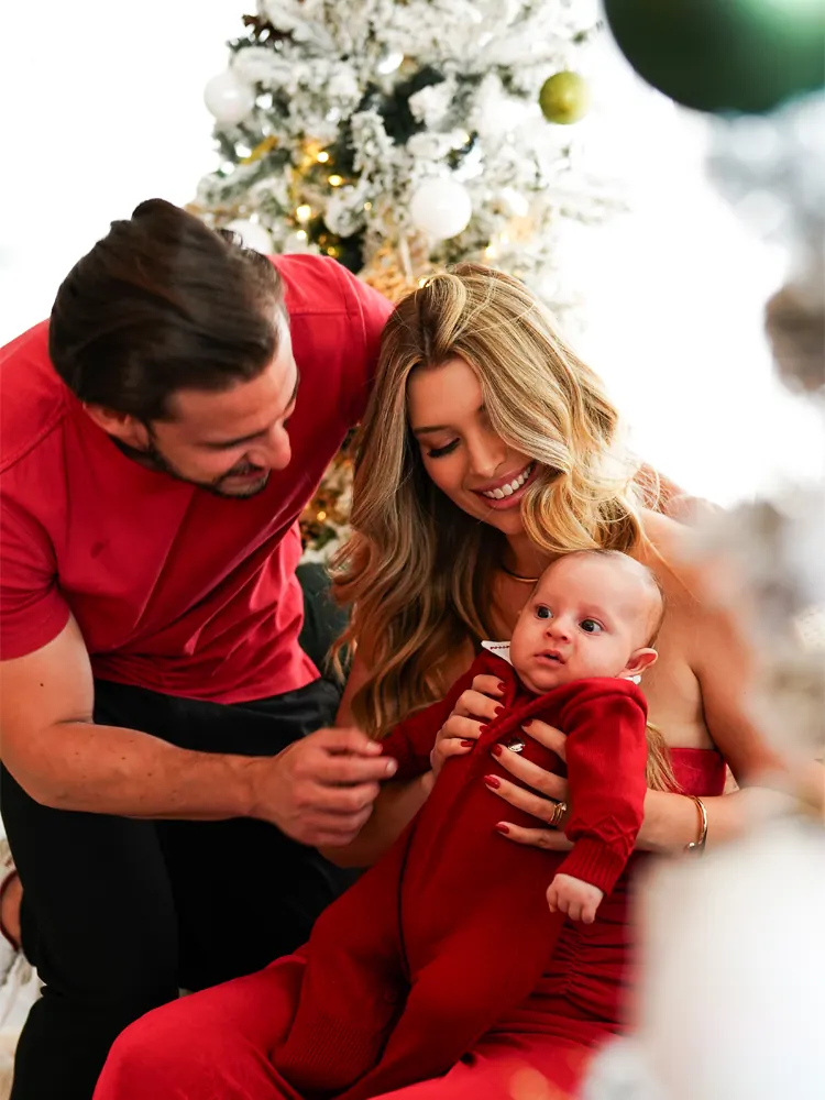 Casal com o bebê no colo, todos de vermelho, diante de árvore de Natal nevada — ensaio de Natal em família no estúdio, Brasília