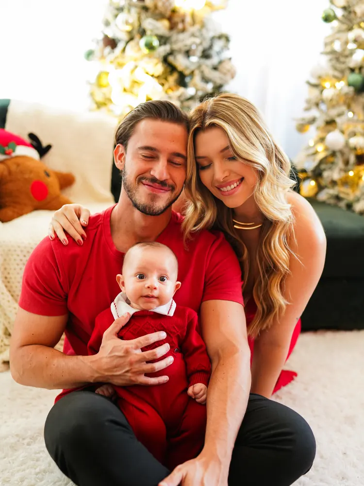 Pai e mãe sentados no tapete com o bebê de macacão vermelho, árvores de Natal ao fundo — ensaio de Natal em Brasília