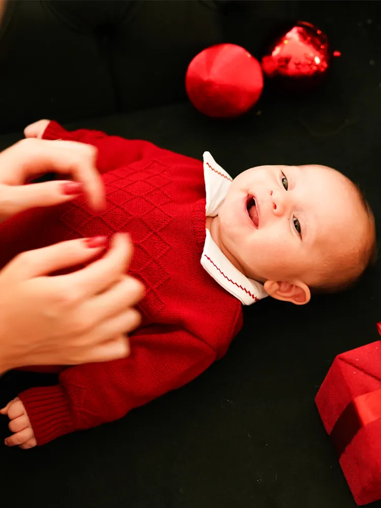 Bebê de macacão vermelho deitado ao lado de bolas de Natal vermelhas — detalhe de ensaio de Natal em Brasília