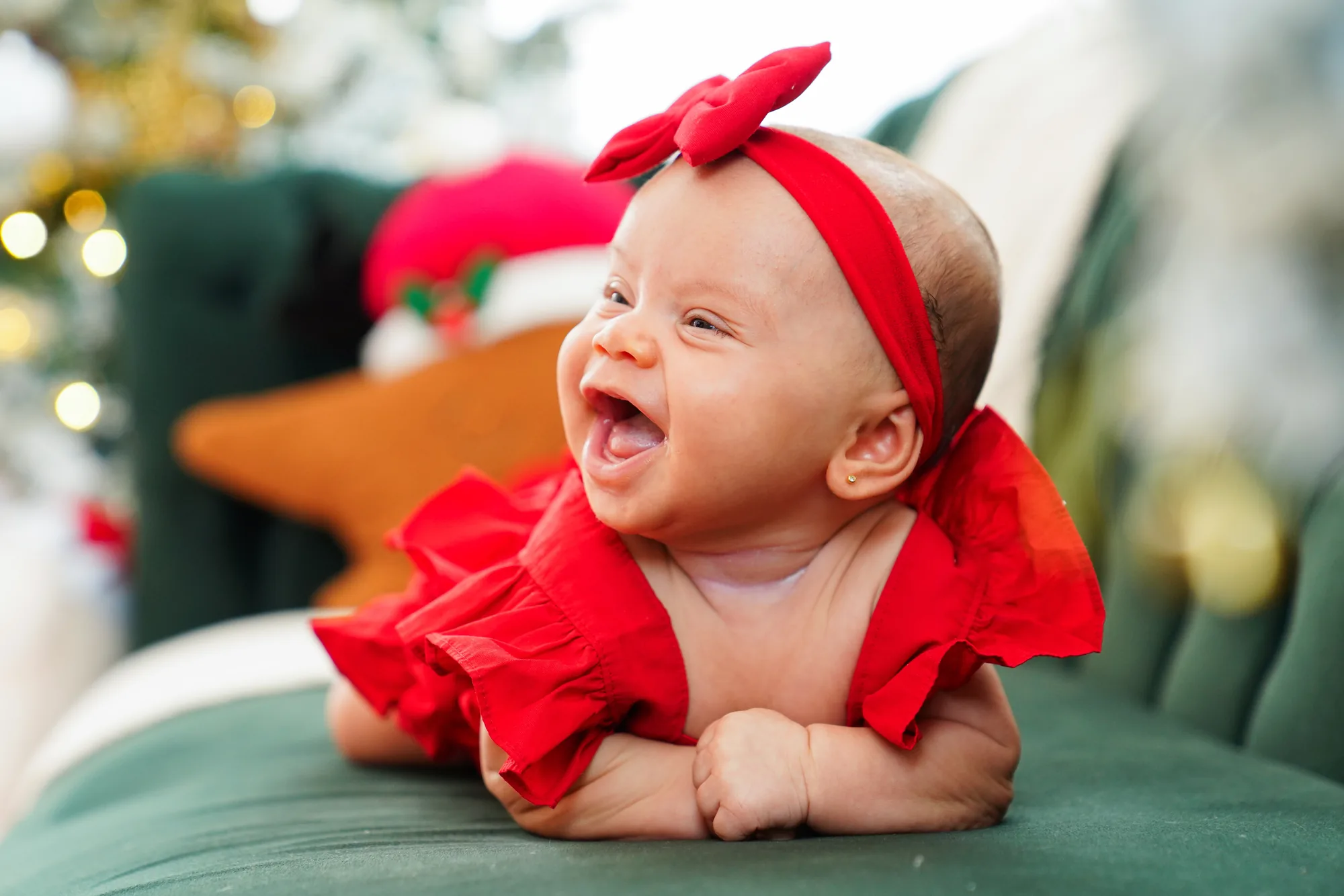 Bebê de laço e vestido vermelho, deitada de bruços em poltrona de veludo verde e sorrindo, árvore de Natal ao fundo — ensaio de Natal em Brasília