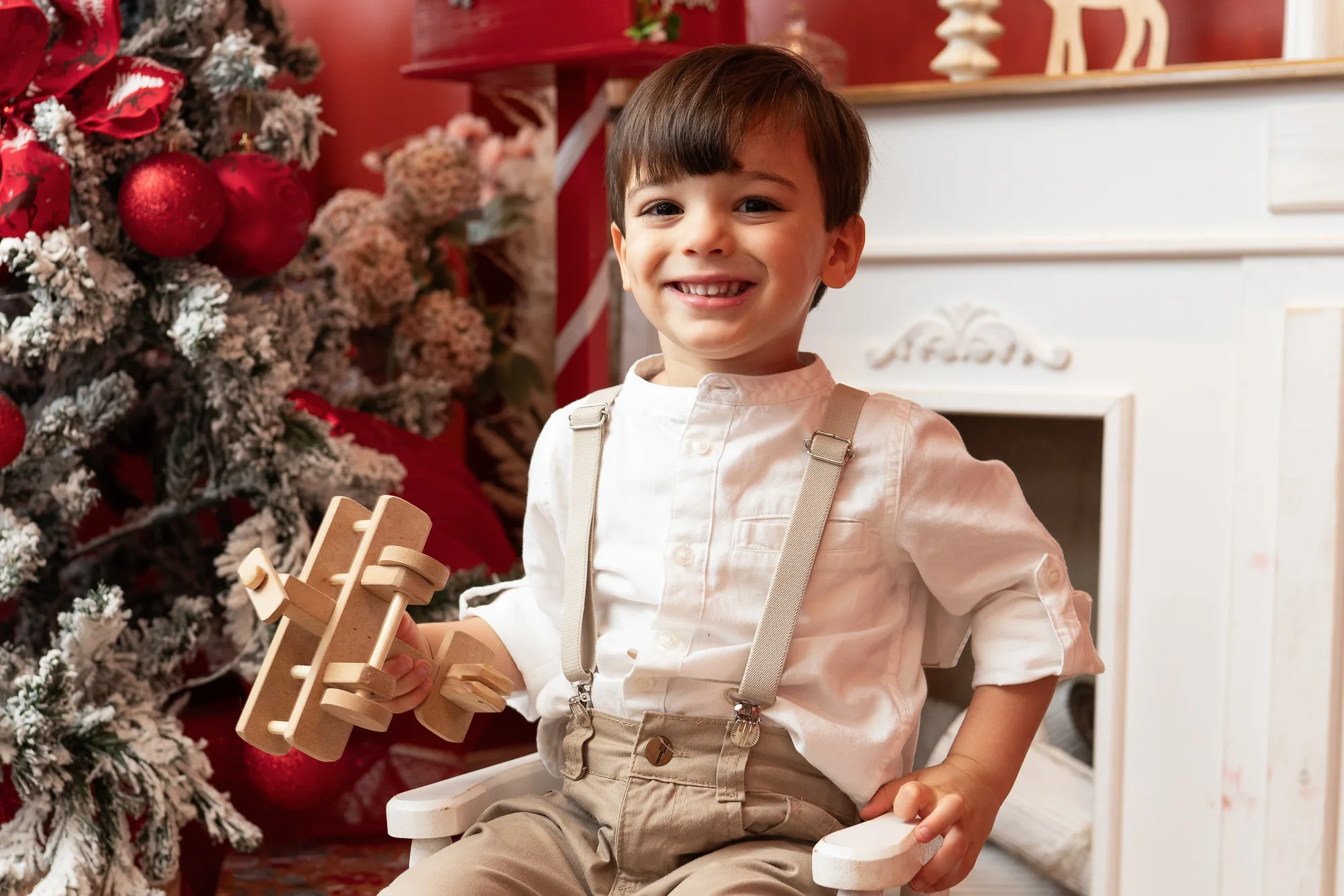 Menino de camisa e suspensórios segurando um aviãozinho de madeira ao lado da árvore de Natal nevada e da lareira branca — ensaio de Natal em estúdio, Brasília