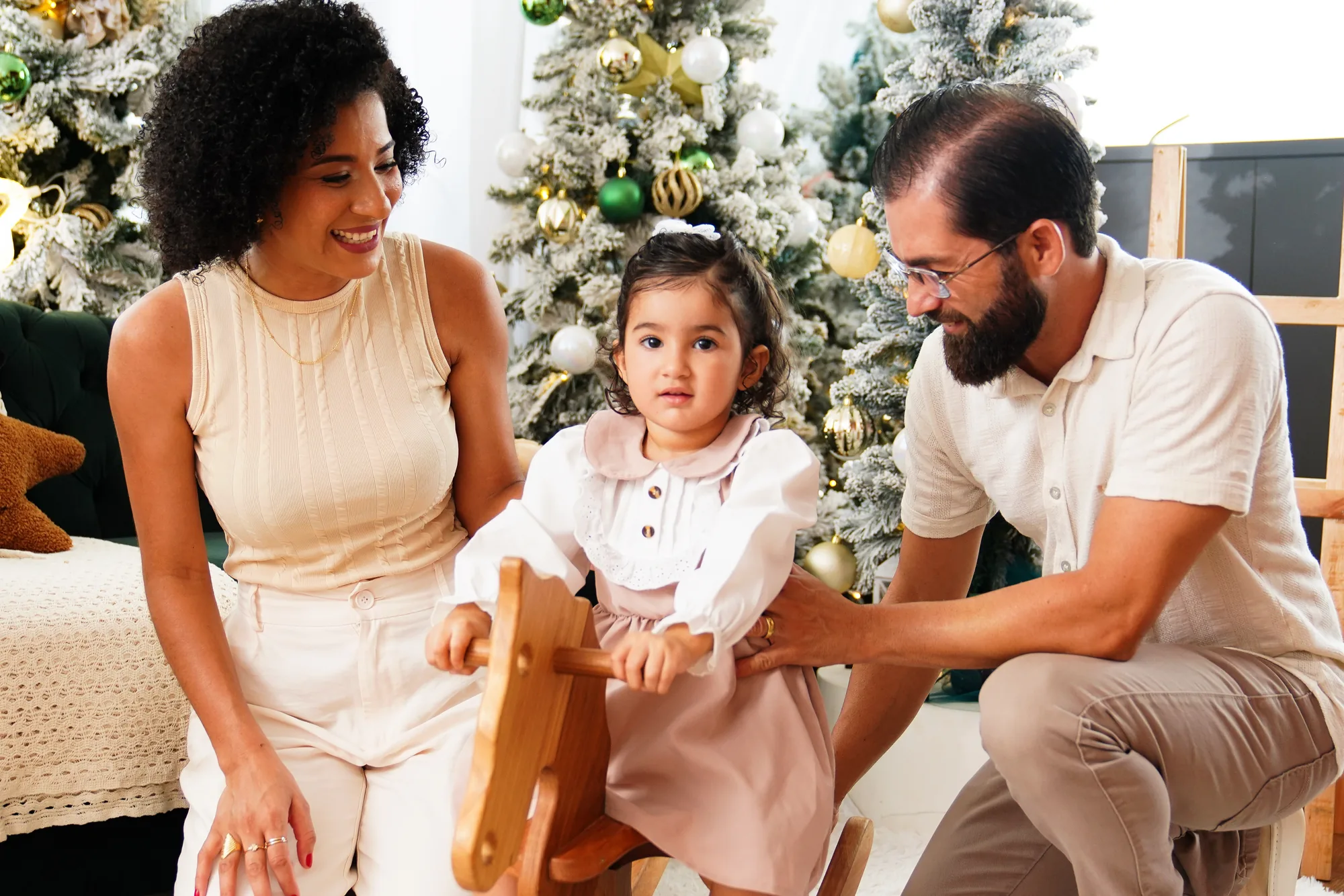 Casal olhando para a filha pequena sentada num cavalinho de madeira, entre árvores de Natal decoradas — ensaio de Natal em família no Lago Sul, Brasília