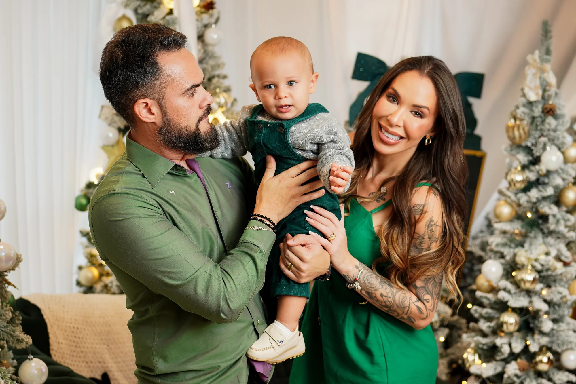 Casal de verde com o bebê no colo entre árvores de Natal douradas e nevadas — ensaio de Natal em família no estúdio do Lago Sul, Brasília