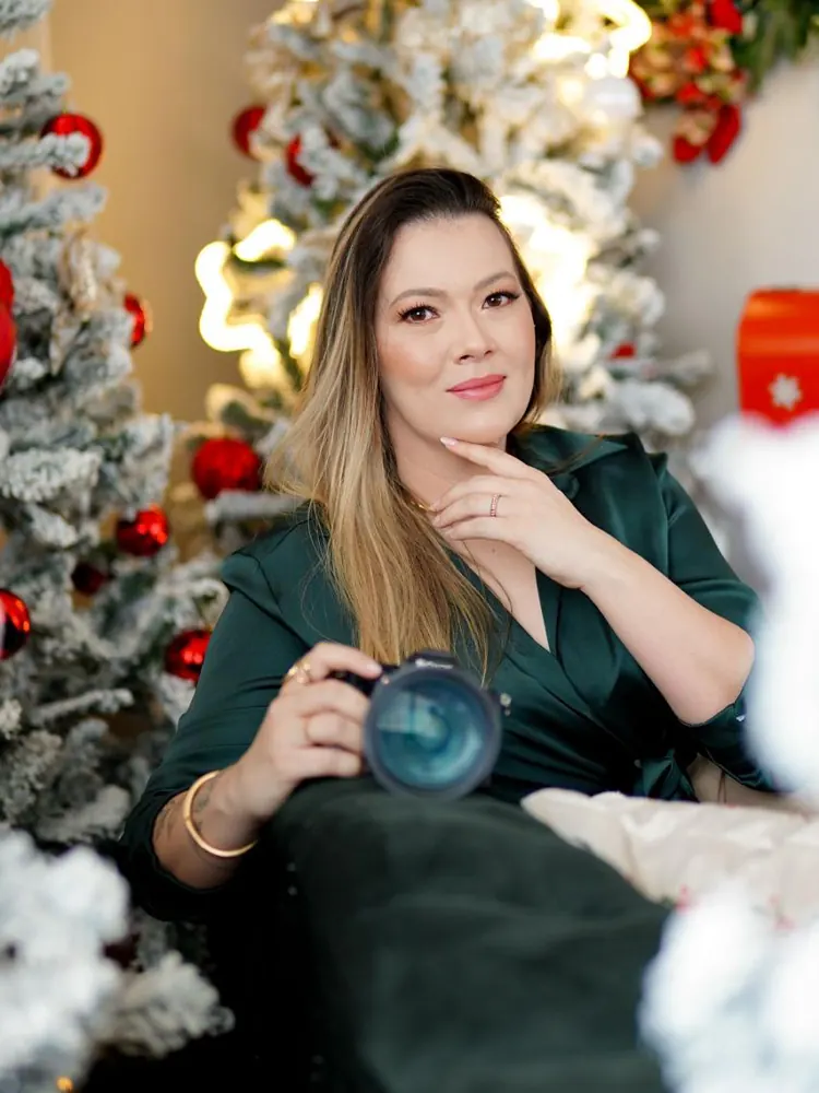 Rebeca Moura de vestido verde, segurando a câmera, entre árvores de Natal nevadas — fotógrafa da RM Foto e Filme, Brasília