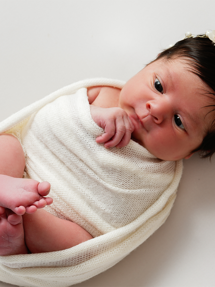 Recém-nascida acordada enrolada em manta de tricô creme, com os pezinhos à mostra — ensaio newborn em estúdio, Brasília