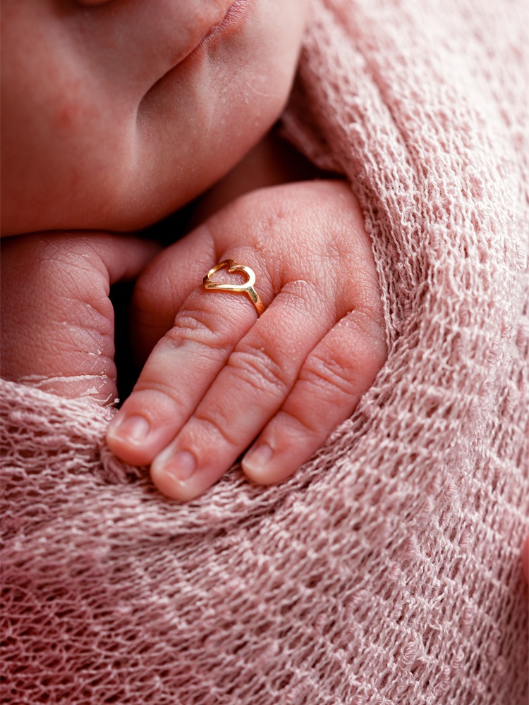 Detalhe da mão do bebê com um anelzinho de coração sobre a manta rosa — ensaio newborn em estúdio, Brasília