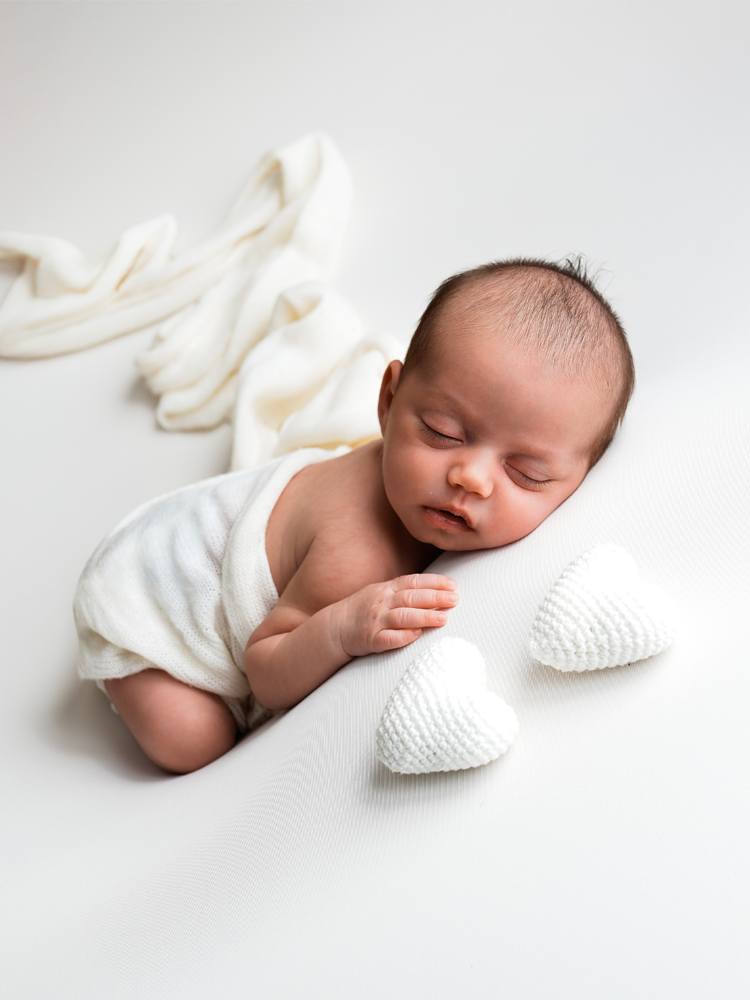 Recém-nascido dormindo de bruços ao lado de dois corações de crochê — ensaio newborn em estúdio no Lago Sul, Brasília