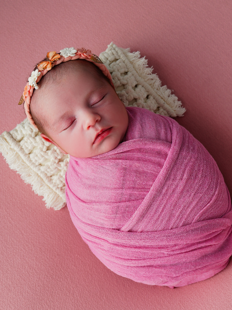 Bebê enrolada em manta rosa com tiara de flores, fundo rosa — ensaio newborn em Brasília