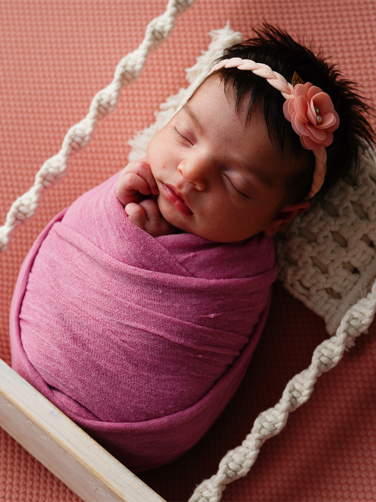 Bebê enrolada em manta lilás com tiara de flor, fundo terracota — ensaio newborn em estúdio, Brasília